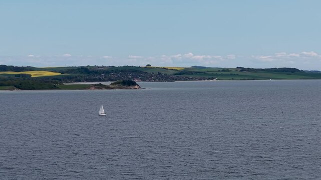Aerial view of a white sailboat cruising on calm water with the rolling hills of Mols Bjerge in the background