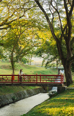 Woman on a red bridge over a stream nature-filled park trees grassy hills tranquil vibes