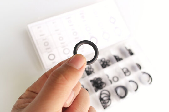A hand holding an O-ring above a clear storage case filled with assorted black rubber gaskets. Perfect for plumbing repair, automotive maintenance, DIY projects, and hardware organization concepts.