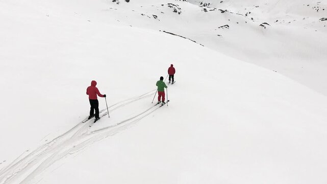 Aerial view of three skiers sliding downhill on pristine untouched snow in the alpine mountains. Red green and black clad skiers descend together leaving clean parallel tracks across the white slope.