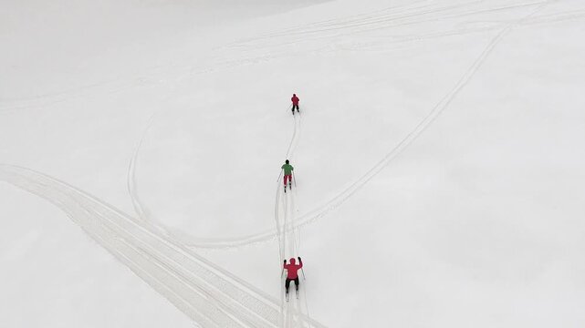 Aerial view of three skiers sliding downhill on pristine untouched snow in the alpine mountains. Red green and black clad skiers descend together leaving clean parallel tracks across the white slope.