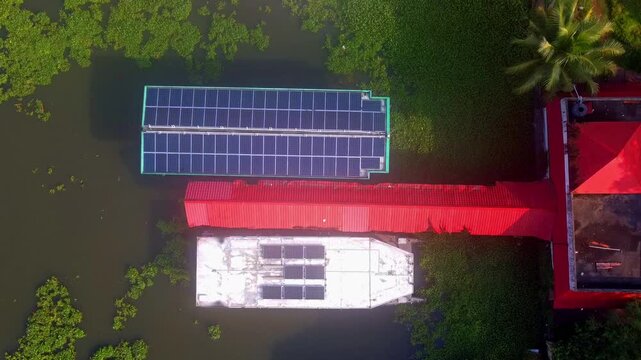 Aerial top-down view of a solar-powered floating ferry or house boat docked in lush green backwaters with solar panels on its roof.