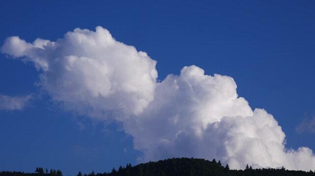 Fluffy, soft, white, cloud moving away in a blue sky behind a green hill with bit speed.