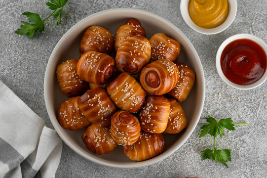 Mini hot dog bites. Sausages baked in a yeast dough cover. Pigs in a blanket in a bowl on a gray concrete background. Top view, copy space.