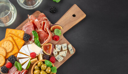 Top view of gourmet charcuterie and cheese board with prosciutto, brie, blue cheese, crackers, figs and berries on dark background with copy space