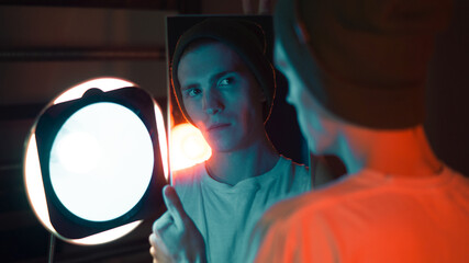 A teenage boy looks into a mirror reflection, wearing a hat, viewed from behind in a studio