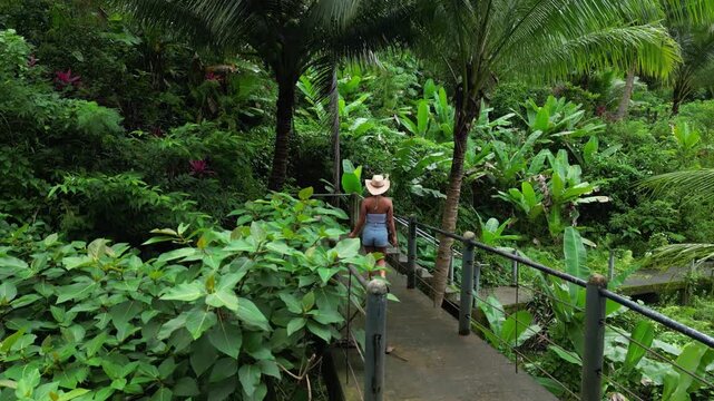 Back-tracking aerial of woman walking elevated metal bridge through dense tropical foliage, gently brushing railings as she moves deeper into the forest in Tanay Rizal, Philippines.