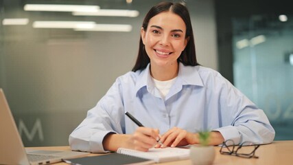 Positive woman diligently works, writing notes with pen in bright office environment, expressing happiness and confidence. Inspires feelings of focus and success in professional setting.