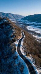 Aerial view of a winding asphalt road descending through snowy hillsides and a valley landscape