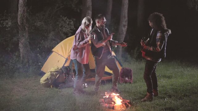 Cheerful diverse friends enjoying a camping trip at night, dancing and singing together around a glowing bonfire while one person plays a small guitar in the middle of a forest clearing