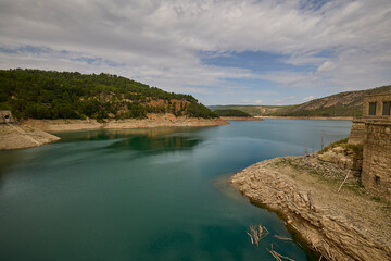 Expansive mountain reservoir with stone dam structures, rocky slopes and green forests under a cloudy sky.