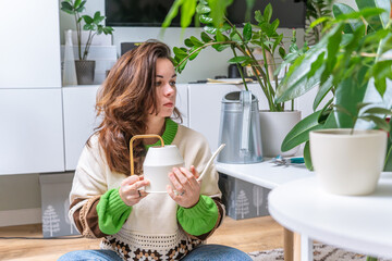 A young woman waters home-grown green plants with a watering can in the living room