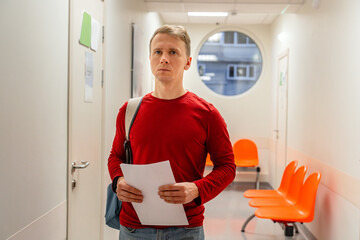 A young man is waiting in the corridor of a medical clinic in front of a doctor's office.