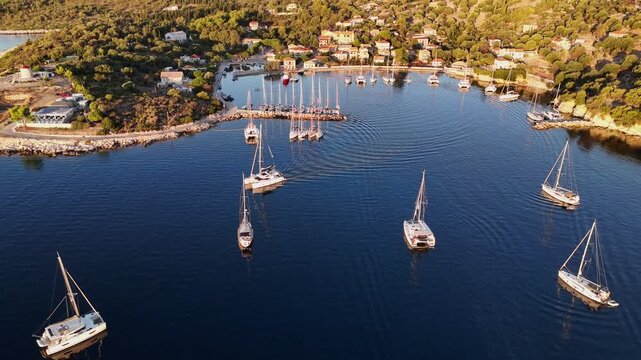 Kastos Island,aerial birds eye view forward towards the harbor on a sunny day.A catamaran is leaving slowly.Lot of boats at the town quay and outside on anchor.Beautiful green Greek Island.