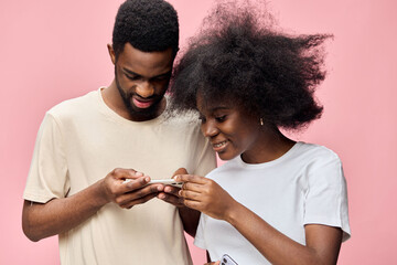 Young African couple enjoying time together, smiling and using a smartphone against a pink background, casual clothing, joyful emotions, modern concept