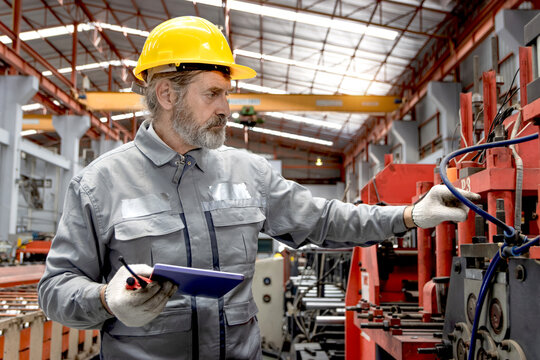 Senior worker wearing safety hard hat, holding tablet and walkie talkie during inspecting machinery in manufacturing factory. Elderly supervisor with helmet working with machine in industrial site. - Powered by Adobe