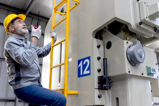 Senior worker wears safety hard hat, holds walkie talkie during climbs a ladder to inspect machine at manufacturing factory. Elderly engineer technician climbing up a ladder at industrial site. - Powered by Adobe