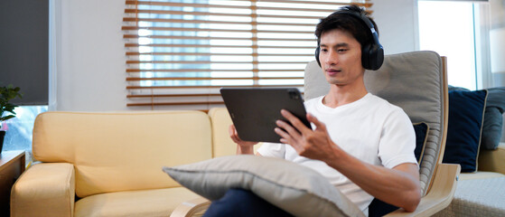 Young Asian man wearing headphones relaxing in cozy living room, smiling while choosing music on tablet