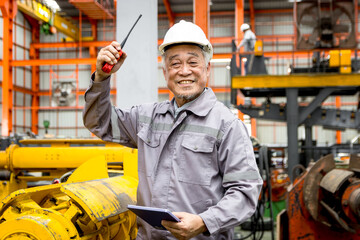 Cheerful Asian senior foreman with safety hard hat holding tablet and walkie talkie during in manufacturing factory. Happy elderly worker with helmet raising hand up to express joy at industrial site.