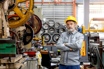 Portrait of senior worker with hard hat standing with arms crossed next to machine at manufacturing factory. Elderly engineer technician with safety helmet working with machinery in industrial site.