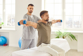 Man patient sitting on the couch in rehabilitation clinic doing sport exercise using dumbbells with nurse. Physiotherapist helping patient in lifting dumbbell. Physical rehabilitation concept.
