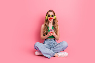 Young stylish woman enjoying music with a smartphone while sitting on a pink background in a casual...