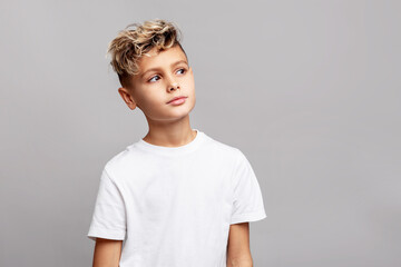 Stylish young boy with trendy modern haircut in studio, wearing white T-shirt and looking aside, isolated on gray background in fashion portrait
