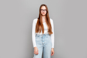 Smart teenage girl student standing in studio, wearing glasses and casual white clothes, confident young female posing on neutral gray background, education and learning theme, indoor portrait
