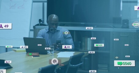 Checking smartphone, mature man sitting in meeting room wearing blue shirt with laptop and overlays