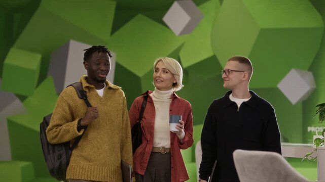 Three diverse colleagues walking side by side, talking and smiling as they entering contemporary office space with bold geometric wall design, one of employees taking seat at desk and opening laptop
