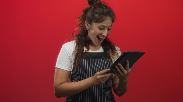 Woman in striped apron and white t shirt tapping and holding a tablet while smiling in a studio red wall setting; digital delight.