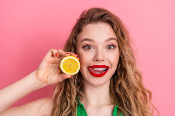 Young woman with braces holding a lemon slice on pink background exuding joy and charm