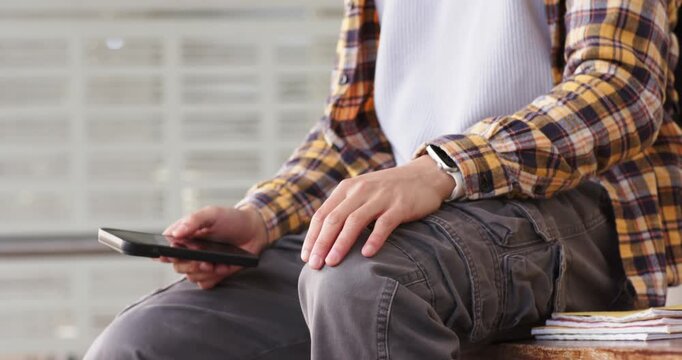 Asian man in 20s tapping smartphone, checking smartwatch, puffing cheeks, waiting at train station