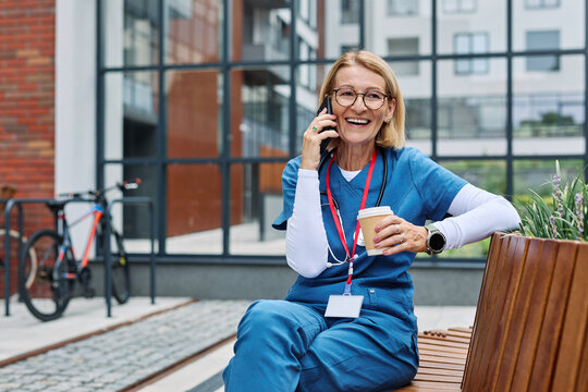 Senior mature woman or a nurse or a doctor during office hours break enjoying relaxing and using mobile phone in a park during a sunny day while smiling and relaxing on a bench - Powered by Adobe