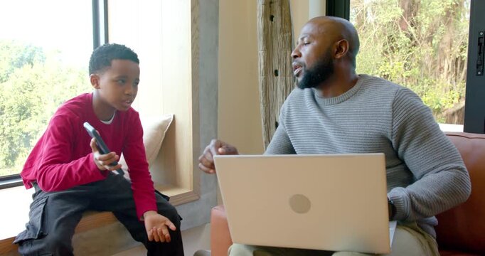 African American father and son pausing laptop after boy offering remote control guiding on sofa