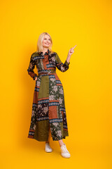 Elegant mature woman in a patchwork dress stands against a bright yellow backdrop and points with confidence for a lifestyle advertisement