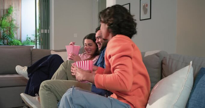 Reacting to shocking TV scene, diverse friends flinching, laughing, sharing popcorn on sofa at home