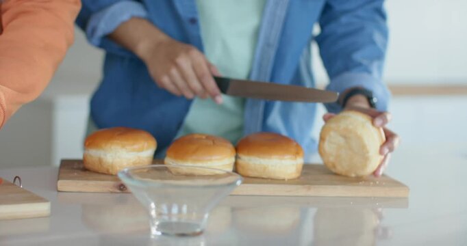 After slicing tomatoes, denim-clad man halving bun with knife at kitchen counter preparing sandwich