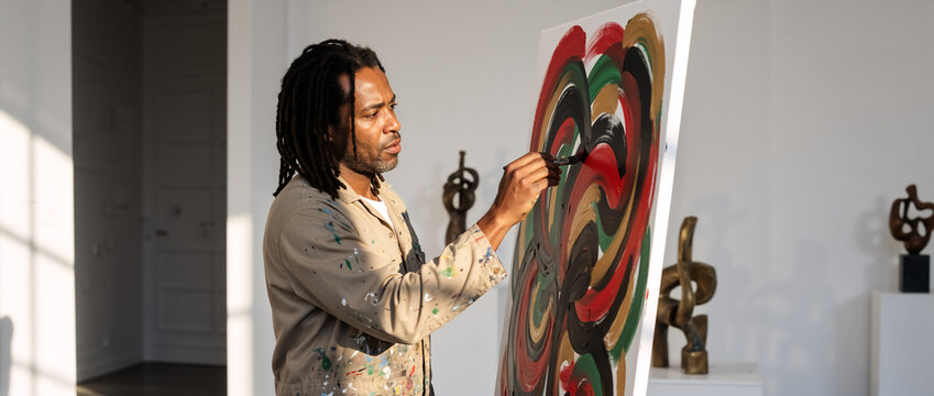 Black artist painting abstract canvas in sunny studio. African American man with dreadlocks creating art with Pan-African colors. Creative painter working during golden hour - Powered by Adobe