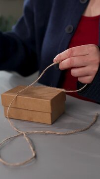 Hands are wrapping a small gift box with string, showing a step-by-step process. This action reflects care and attention to detail in preparing a nice surprise for Valentine's Day.