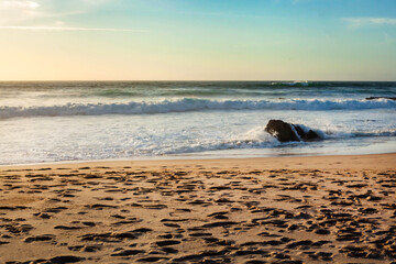 Beach at sunset with glowing sky, waves crashing against the shore and a single dark rock in the surf, creating a calm yet dramatic coastal mood.