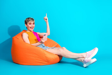 Young woman relaxing on bright orange beanbag in a colorful studio using a smartphone and pointing up with a cheerful expression