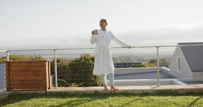 Mid adult Asian woman leaning on terrace railing at sunrise sipping mug, scanning hillside for calm