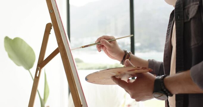 After dipping brush in red paint, painter's hands are outlining canvas on easel finishing border