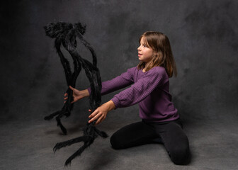 Small girl sitting on floor and gently petting large black toy spider against dark background. Ideal for kid themes, imagination, and creative visuals.