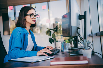 Young professional woman programmer sits at a modern office desk working on a computer monitor with sticky notes and a plant