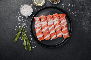 Raw minced meat rolls wrapped in bacon on black plate with spices