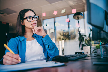 Thoughtful professional woman at a desk planning and taking notes in a modern office with computer monitor and sticky notes