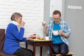 Woman looks into the teapot while drinking tea with older woman, embodying care and leisure time between generations, concept of senior living and support.