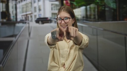 Woman with glasses smiling while pointing finger to camera in urban street beside building walkway railing; confidence.